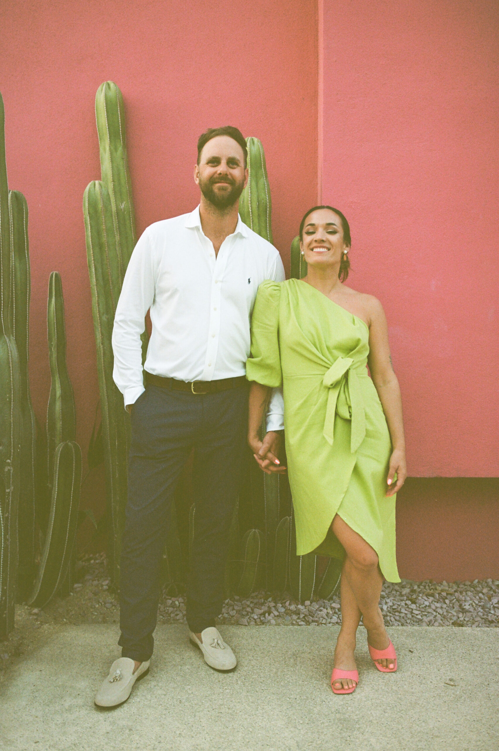 sidewalk couple photo shoot against pink wall with cacti in background. Downtown San Jose Del Cabo, Mexico. white button up with brown belt, blue slacks and light tan shoes. Chartreuse green one shoulder quarter sleeve wrap dress that ties in the front. Bright coral colored kitten heels from Target. colorful wedding portraits. pink lipstick slicked back up-do hairstyle. Natural makeup. Colorful couple portraits. desert landscaping. 