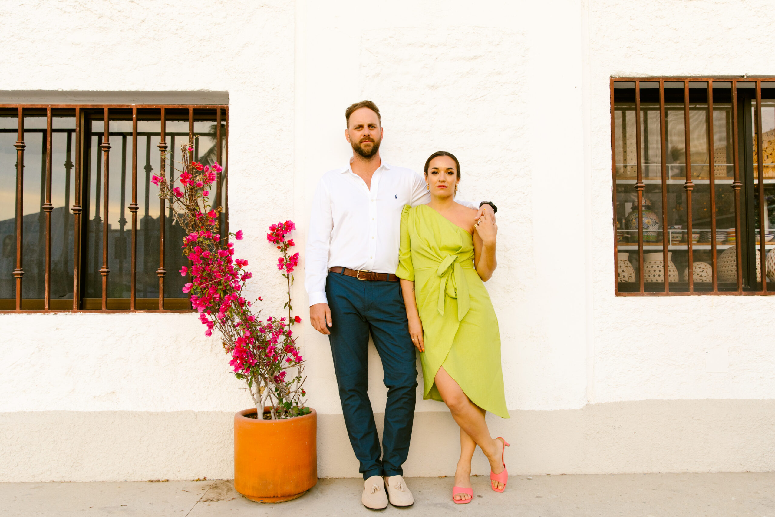 sidewalk couple photo shoot against white and tan wall next to fuchsia florals in a clay peach pot. Downtown San Jose Del Cabo, Mexico. white button up with brown belt, blue slacks and light tan shoes. Chartreuse green one shoulder quarter sleeve wrap dress that ties in the front. Bright coral colored kitten heels from Target. colorful wedding portraits. red lipstick slicked back up-do hairstyle. 