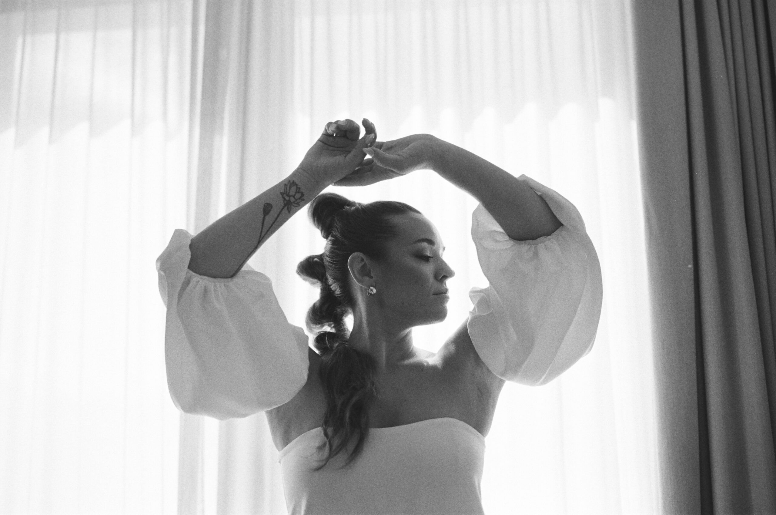 black and white photo of bride with arms in the air floral tattoo artsy wedding portrait verona gown kamperett in front of backlit window mohawk high bubble ponytail hairstyle unique wedding hair edgy wedding hair