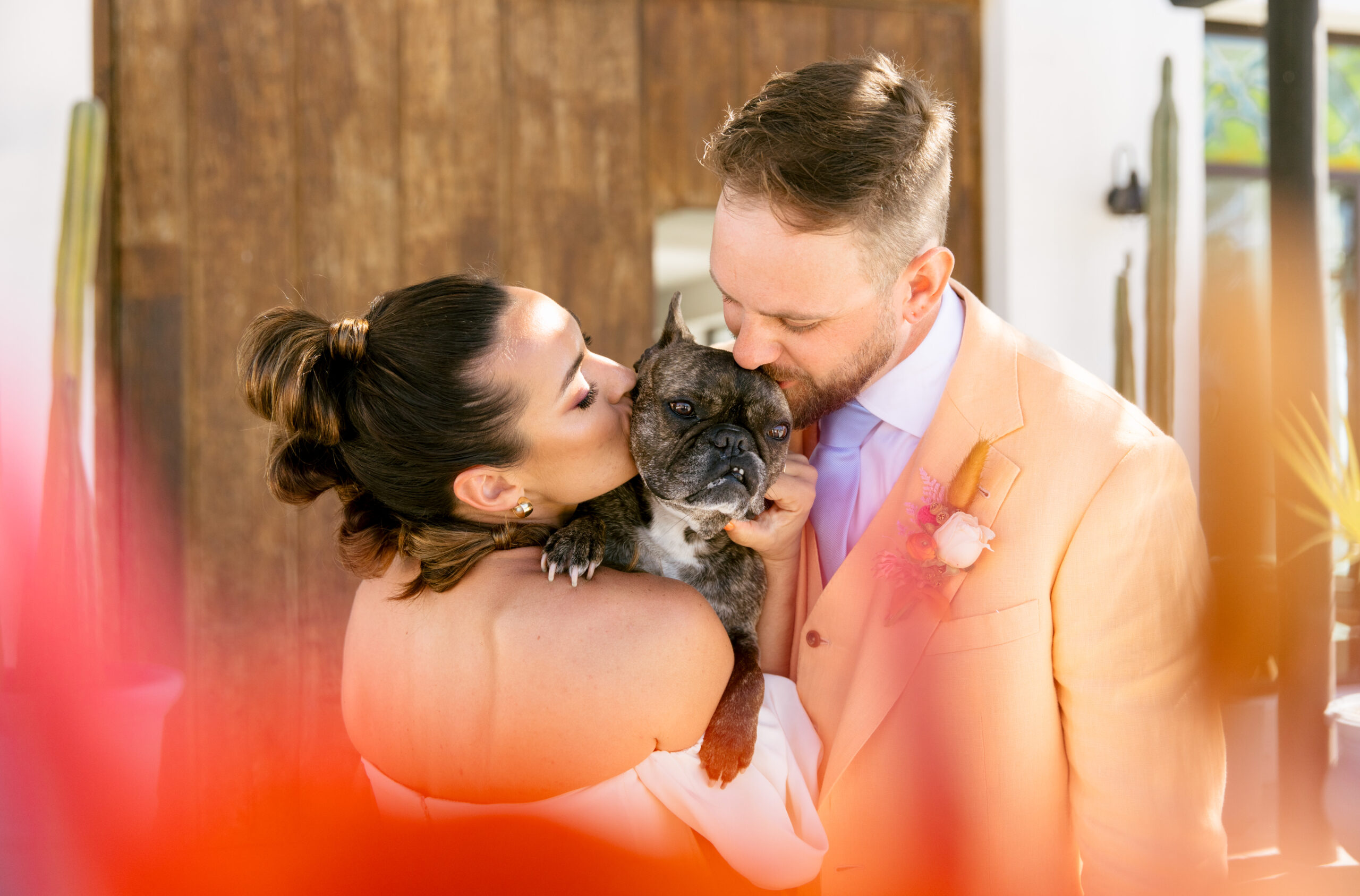 verona wedding gown kamperett strapless wedding dress with detachable sleeves. colorful wedding in mexico. bridal wedding portrait mohawk high bubble ponytail hairstyle. chartreuse dolce vida shoes. 
groom peach cotswold tailors suit with light blue tie and orange and pink boutenir. bride and groom wedding portrait holding brindle french bulldog with hot pink out of focus floral in the foreground. artsy wedding portraits. 