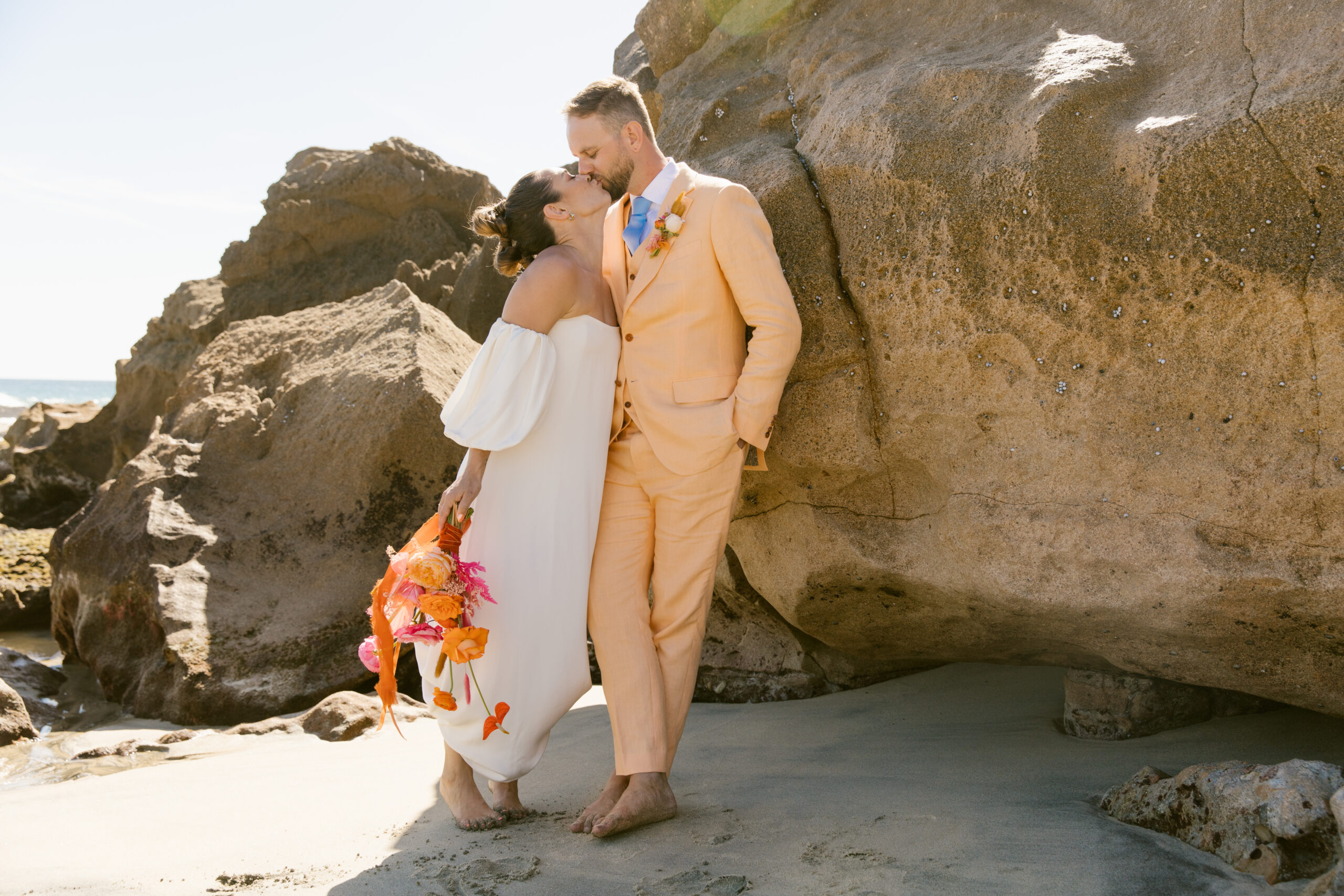 verona wedding gown kamperett strapless wedding dress with detachable sleeves. colorful pink and orange wedding bouquet. colorful wedding in mexico. bridal wedding portrait mohawk high bubble ponytail hairstyle. chartreuse dolce vida shoes. 
groom peach cotswold tailors suit with light blue tie and orange and pink boutenir. bride and groom wedding portraits kissing on beautiful beach in mexico artsy portraits