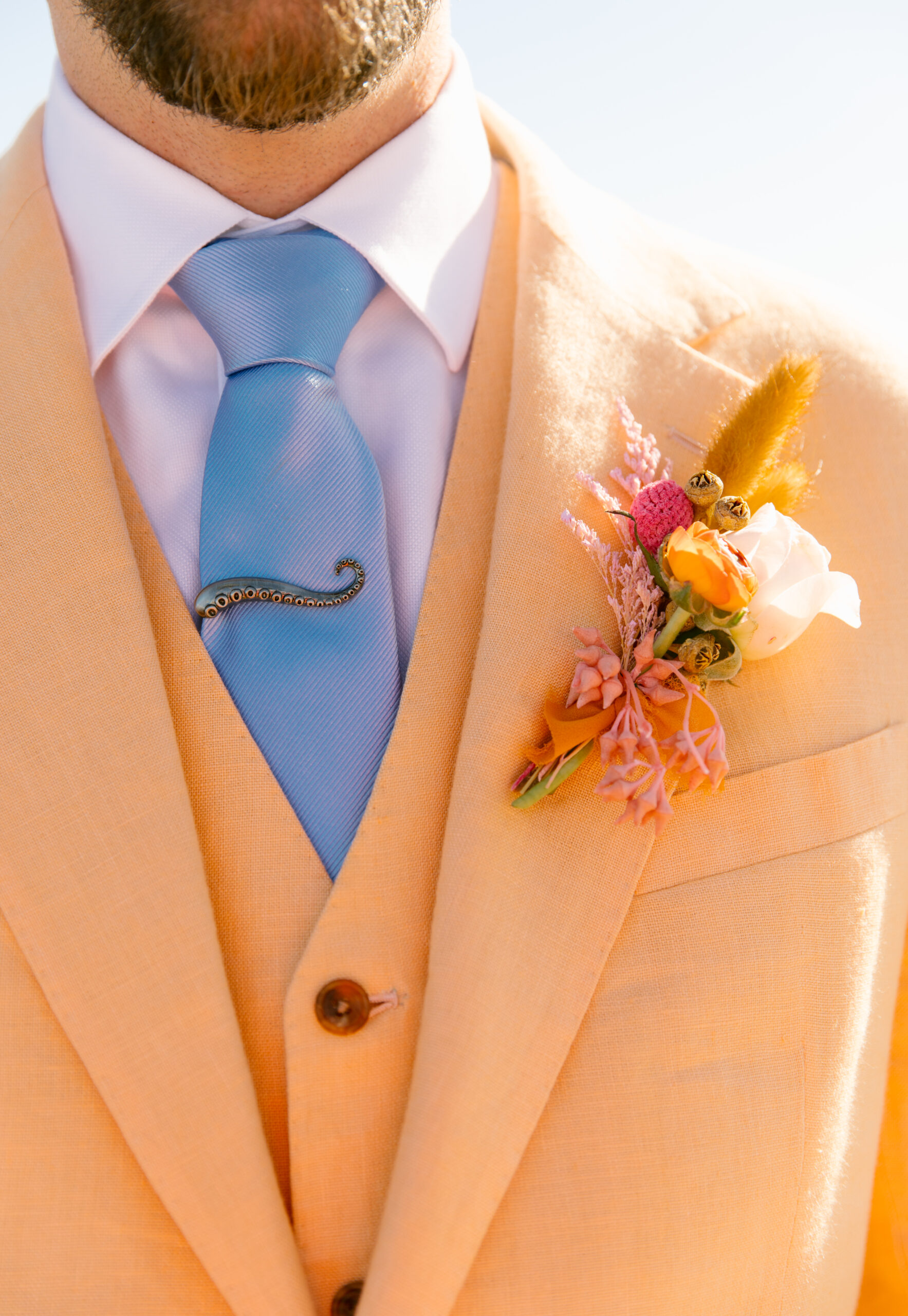 bright peach or coral linen suit cotswold tailors with pink orange and white boutenir. light blue tie with octopus tentacle tie clip. colorful groom style. 