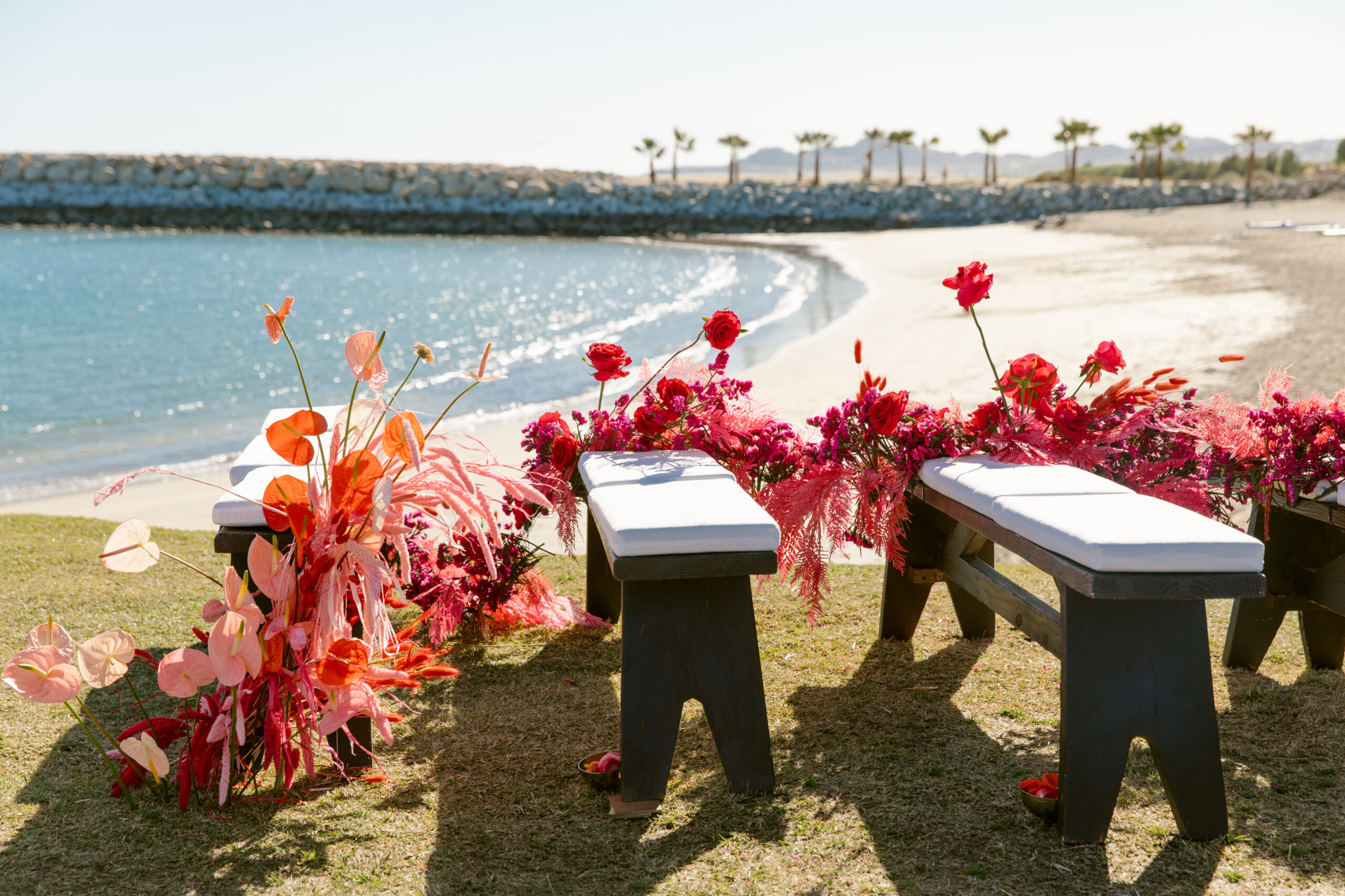 wedding florals benches unique and colorful wedding ceremony florals. wild florals wrapped through benches for mexico wedding ceremony on jetti at beach club. dark brown wooden benches with white cushions. pink and peach and orange and yellow colored florals. looks like florals are growing out of the benches. birds of paradise dried florals roses. lilies ocean beach view in the background. palm trees white sand beaches. blue clear ocean water. blue skies. 
