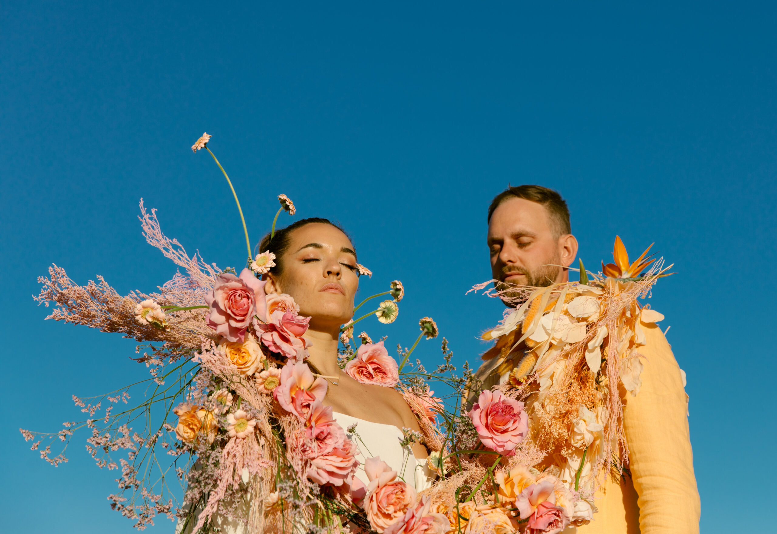 close up vogue inspired wedding portraits. bride and groom wrapped in colorful florals pinks oranges yellows dried florals roses daisies blue skies. light orange linen groom suit
