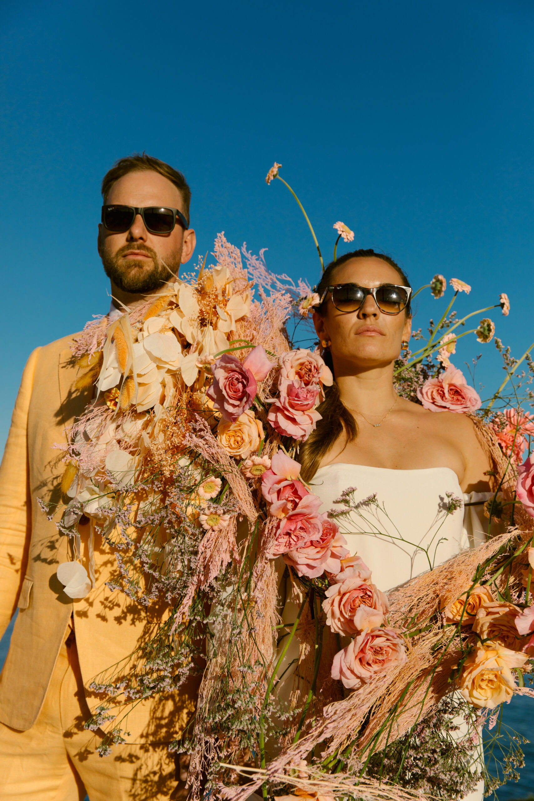 close up vogue inspired wedding portraits. bride and groom wrapped in colorful florals pinks oranges yellows dried florals roses daisies blue skies. verona gown strapless wedding dress kamperett. light orange linen groom suit cotswold tailors. bride and groom sunglasses portrait