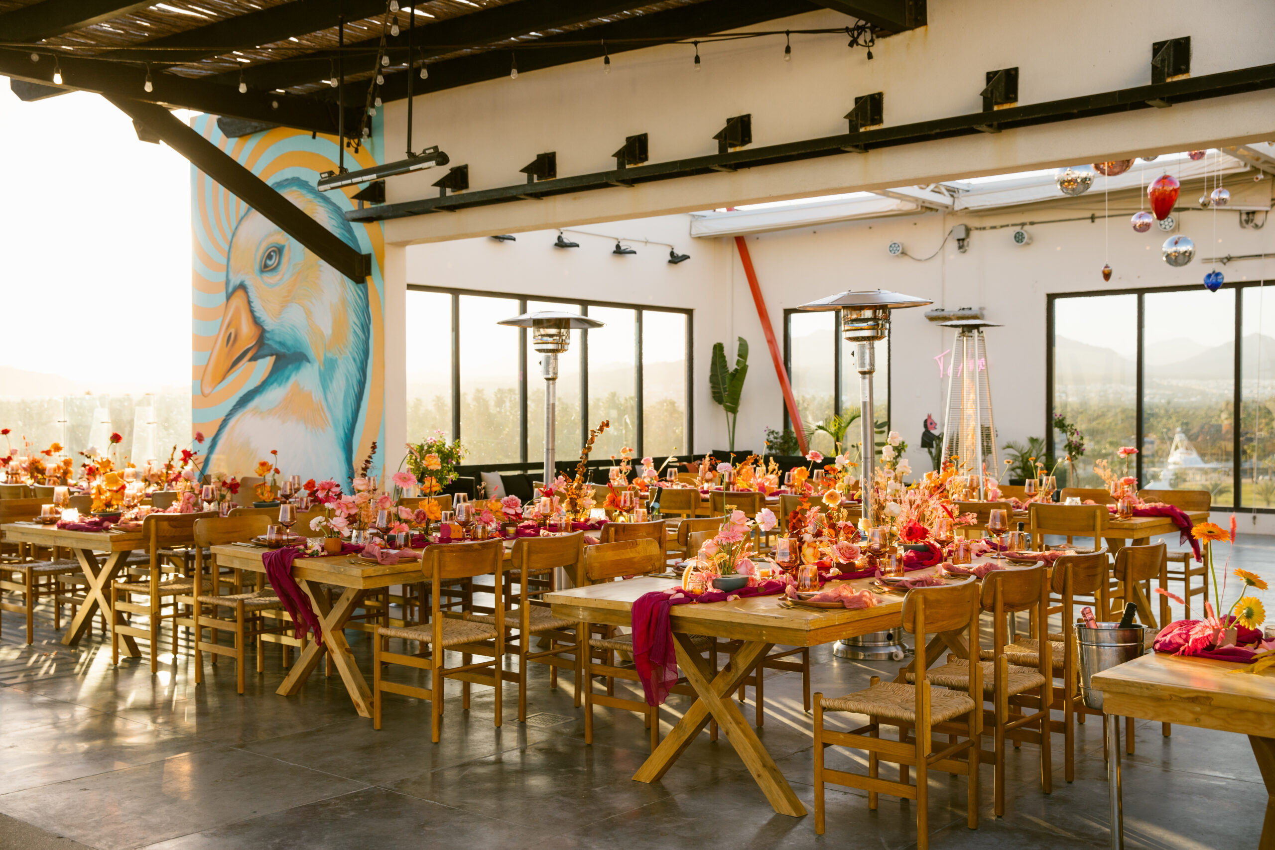 hotel el ganzo rooftop. colorful mexican mural. colorful wedding florals. bright pinks, red, orange, yellow. fuchsia gauze runner on light wooden table with wooden chairs. smokey pink colored glassware. wedding reception tabletop. 
orange and pink custom table number signs. 
blue skies. ocean view. rooftop wedding dinner reception. mauve clay handmade plates. gold flatware

hanging blown glass hearts and disco balls. te amo neon sign. 