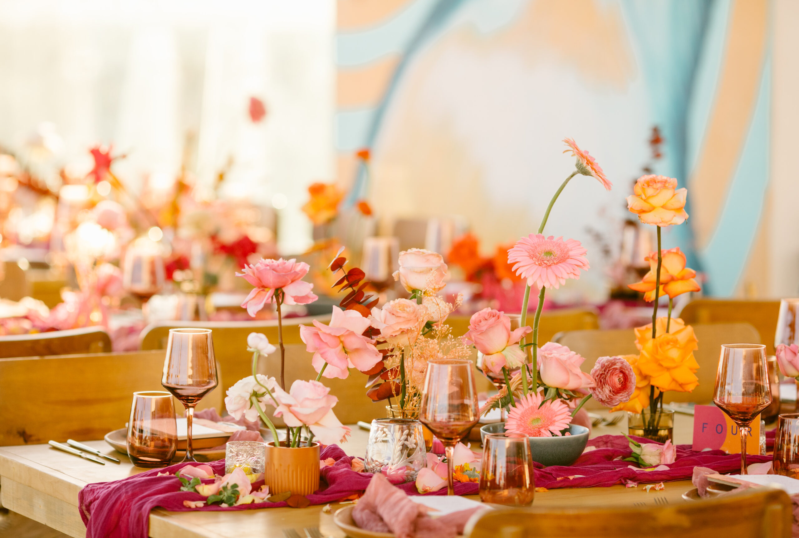 hotel el ganzo rooftop. mexican mural in background of wedding reception. colorful wedding florals. bright pinks, red, orange, yellow. fuchsia gauze runner on light wooden table. smokey pink colored glassware. wedding reception tabletop