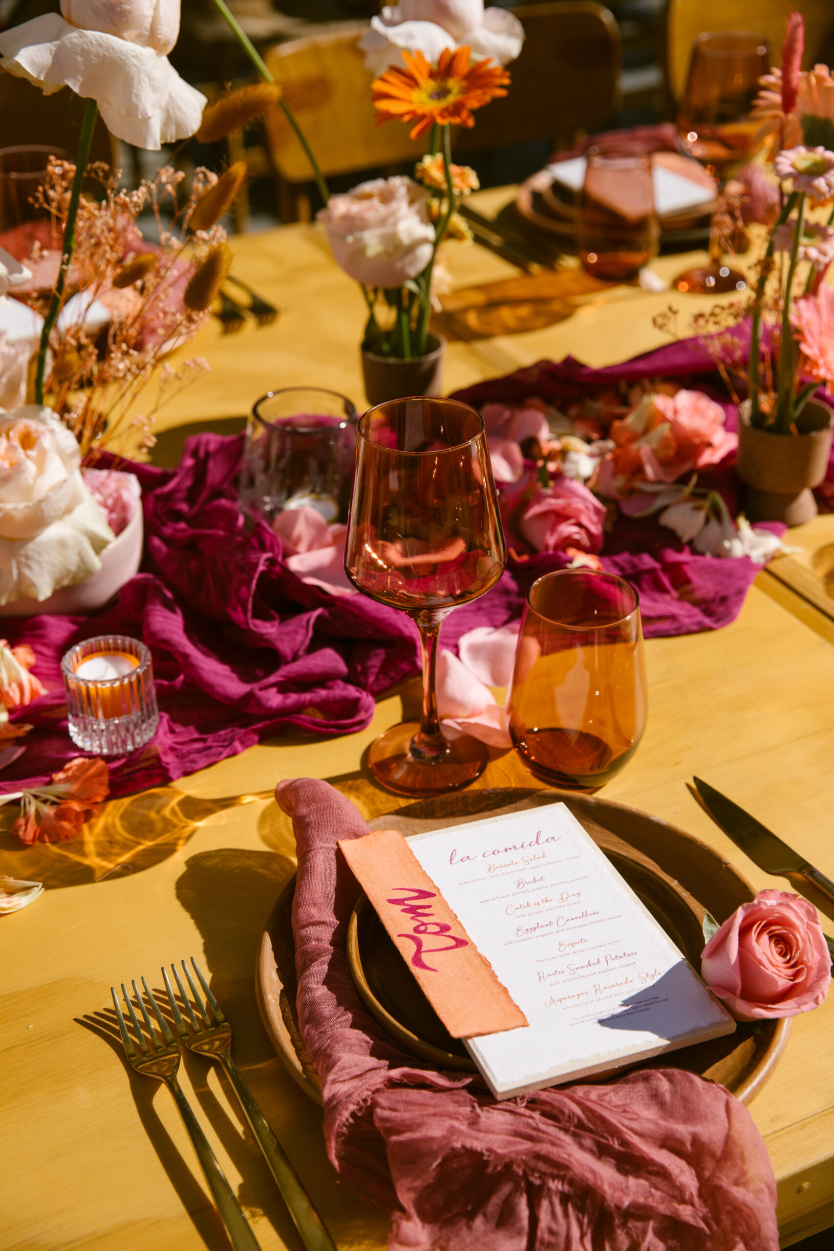 hotel el ganzo rooftop. colorful wedding florals. bright pinks, red, orange, yellow. fuchsia gauze runner on light wooden table with wooden chairs. smokey pink colored glassware. wedding reception tabletop. 
orange and pink custom table number signs.  mauve clay handmade plates. gold flatware. dusty rose pink gauze napkin. small clear pleated candle votives with colored candles. textured paper white menu with purple orange and pink writing on a light wood board with hand painted peach and pink nametag
