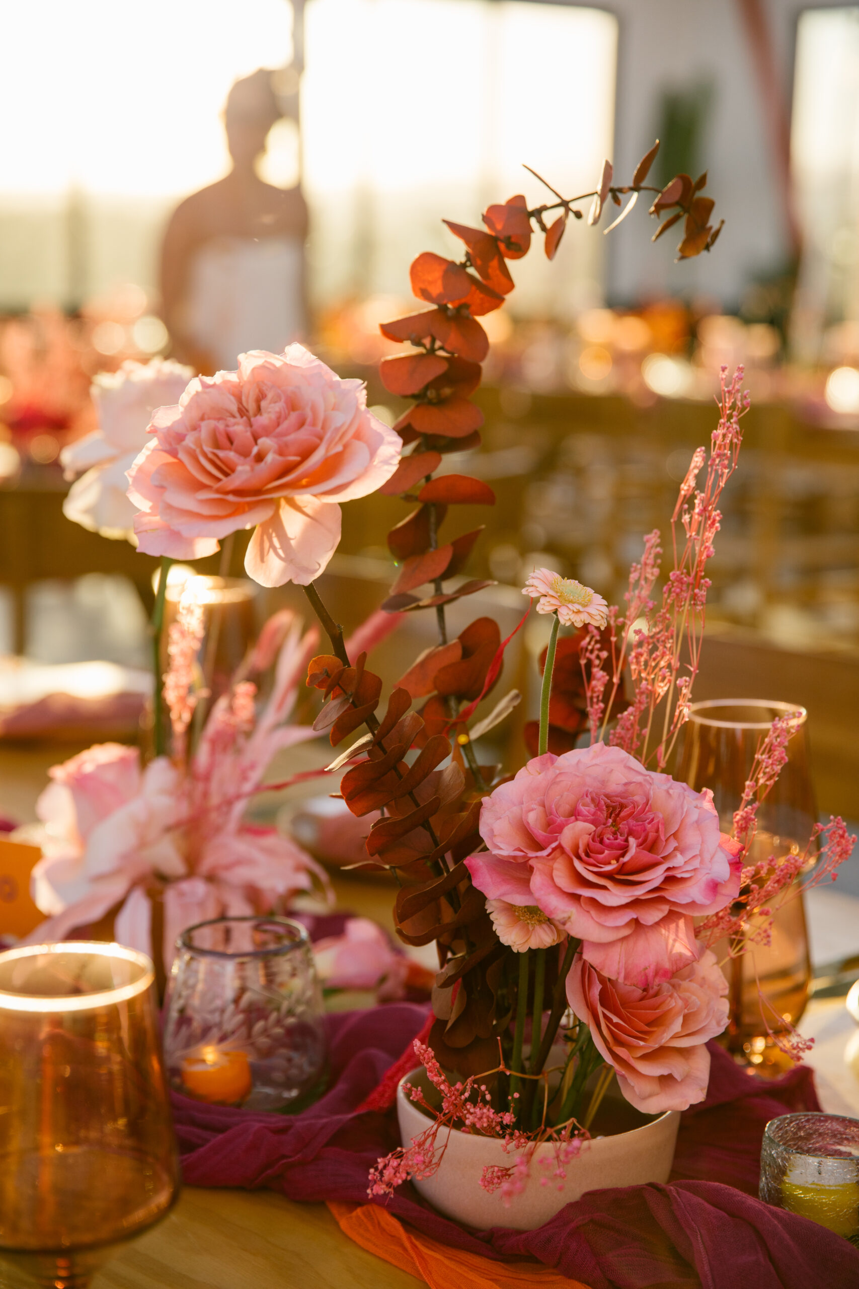hotel el ganzo rooftop. colorful wedding florals. bright pinks, red, orange, yellow. fuchsia gauze runner on light wooden table with wooden chairs. smokey pink colored glassware. wedding reception tabletop. 
orange and pink custom table number signs.  mauve clay handmade plates. gold flatware. dusty rose pink gauze napkin. small clear pleated candle votives with colored candles. bride out of focus in background.
