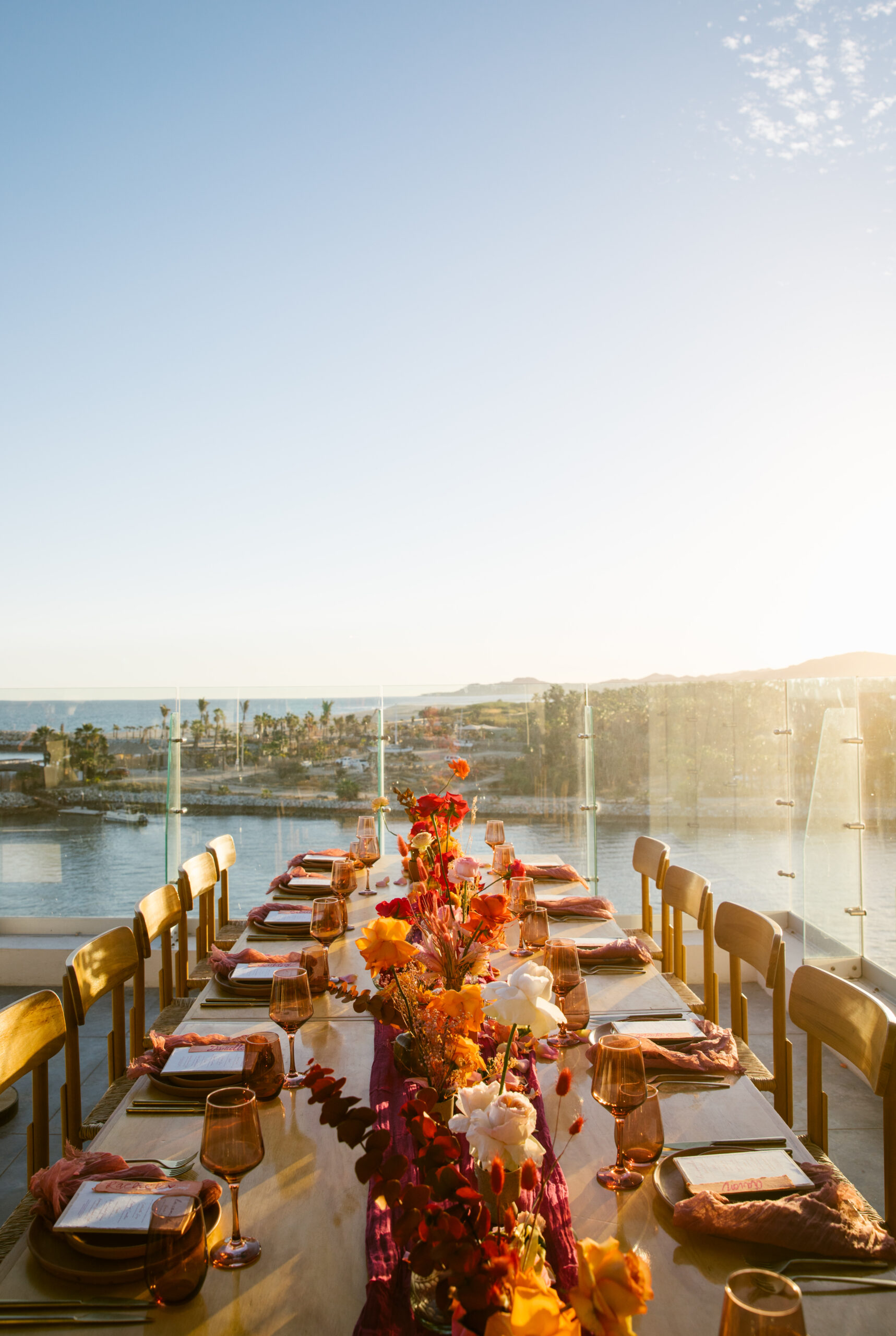 hotel el ganzo rooftop. colorful wedding florals. bright pinks, red, orange, yellow. fuchsia gauze runner on light wooden table with wooden chairs. smokey pink colored glassware. wedding reception tabletop. 
orange and pink custom table number signs. 
blue skies. ocean view. rooftop wedding dinner reception. mauve clay handmade plates. gold flatware