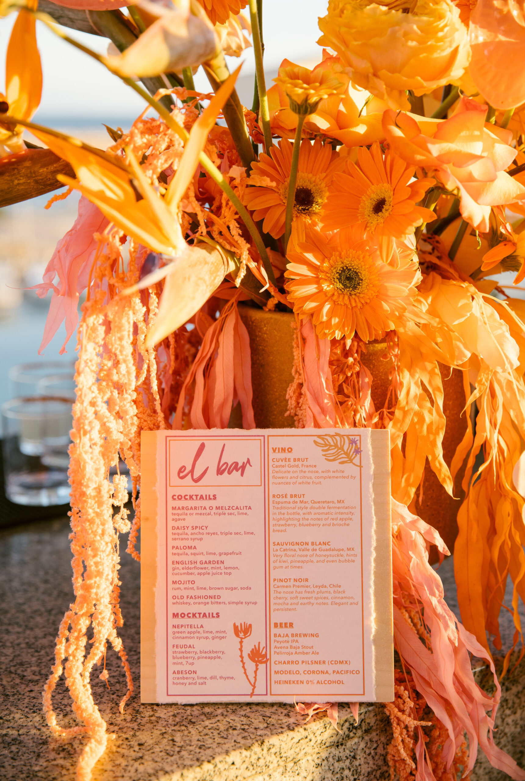 bright orange daisy rose birds of paradise wedding bar arrangement. 
bar menu with pink orange and purple on white recycled paper on light wood board. custom colorful wedding bar menu