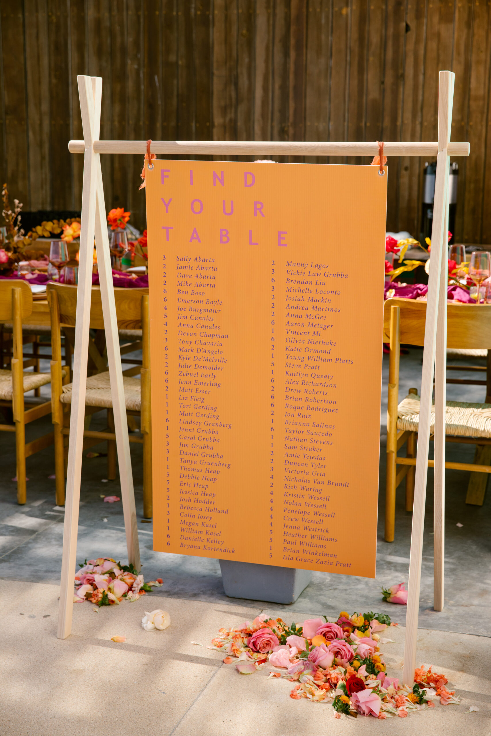 orange and pink and purple wedding seating chart on light wooden arbor. flower heads decorated around seating chart on the floor. hotel el ganzo rooftop. colorful wedding florals. bright pinks, red, orange, yellow. fuchsia gauze runner on light wooden table with wooden chairs. smokey pink colored glassware. wedding reception tabletop. 
orange and pink custom table number signs. 

