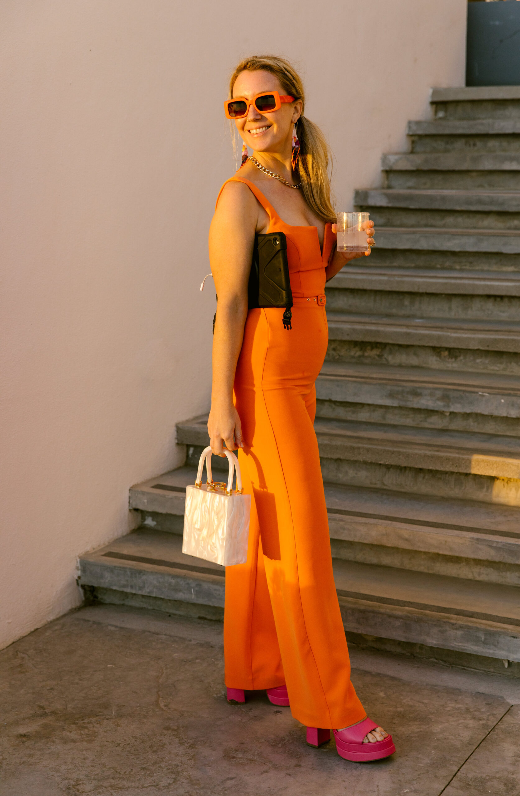 monochromatic orange look with orange jumpsuit and orange sunglasses fuchsia pink chunky heels and a pearl box purse with chunky gold chain necklace. mexico destination wedding guest colorful outfit