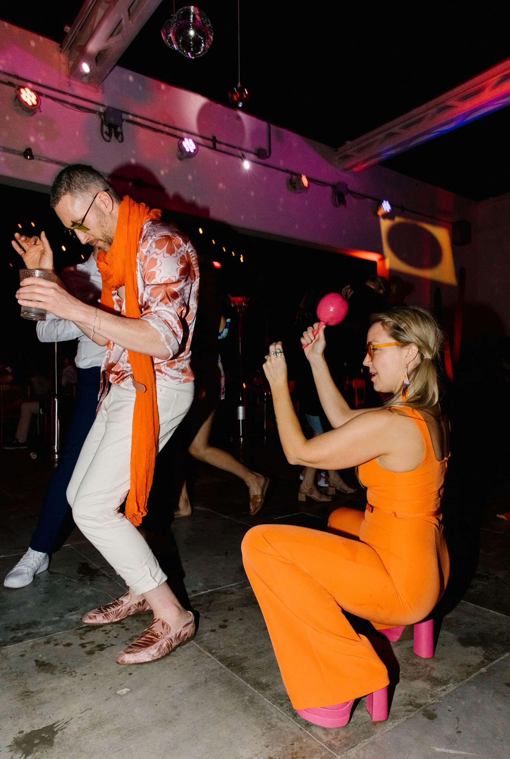disco ball and hanging blown glass heart ceiling installation wedding dance party moody colorful lighting. colorful wedding guest outfits. orange jumpsuit with chunky fuchsia heels. velvet pink boat shoes