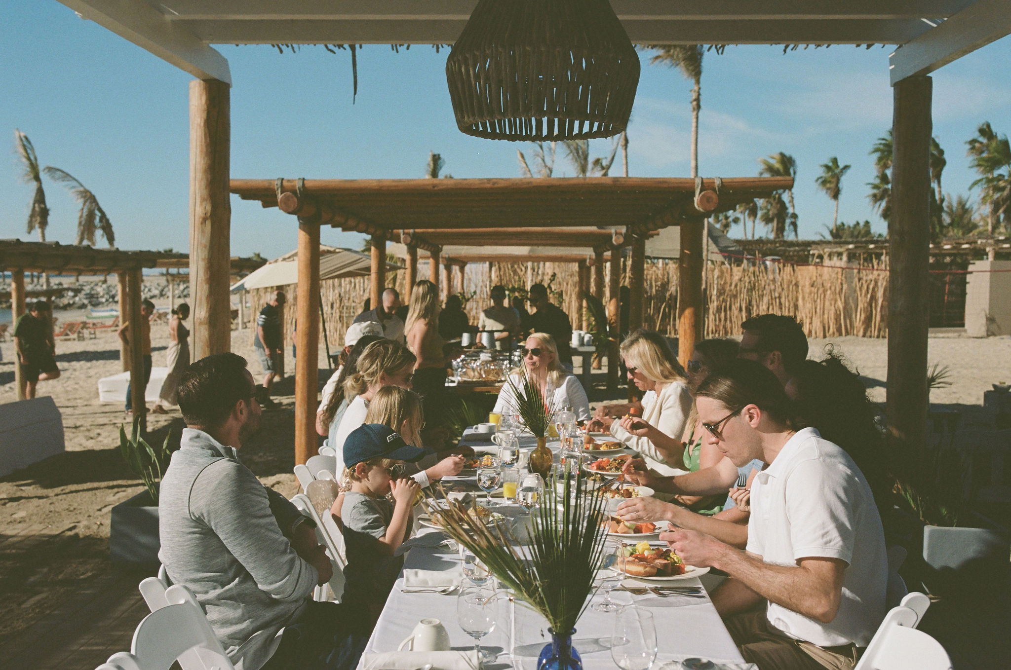 film style photo with grainy filter. 
al fresco style breakfast on the beach. Long tables with basket chandeliers under wooden auning. palm leaf decor on tables. family eating together. 