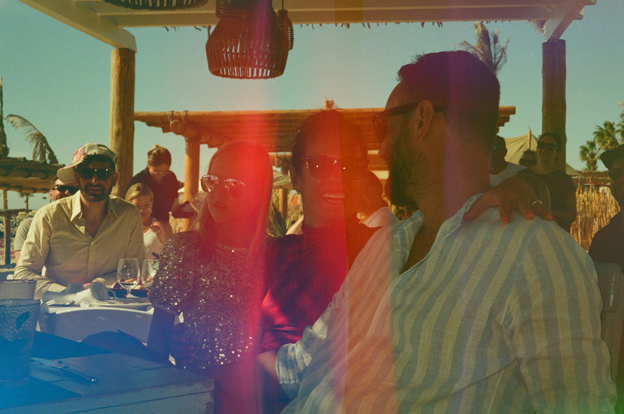 film style photo with sun flare and fuzzy filter. Outdoors man and woman sitting down at table looking at each other and smiling. Man wearing white and blue striped shirt. woman has arm around man. Coral nails. Pink and purple tight turtle nexk dress with slit. Little girl in background with sparkly shirt. Man sitting at another table in the background wearing a hat to the side. Al fresco daytime dining under auning with basket chandelier on the beach.