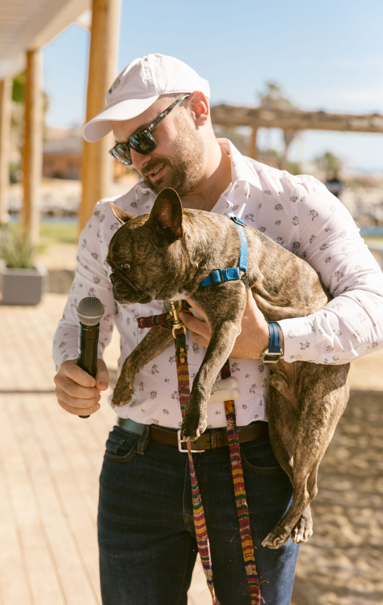 man with denim jeans, sunglasses, white hat and white long sleeve button up holding brindle french bulldog in one hand, and holding a wireless microphone up to the dog in another hand. outdoors under wooden auning with beach in the background. 