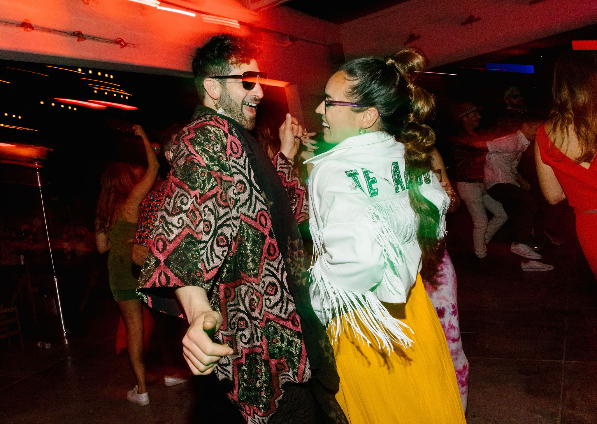 blurry photo style te amo bridal jacket with white fringe yellow after party dress high bubble pony tail man with pink and green colorful cloack. dance party