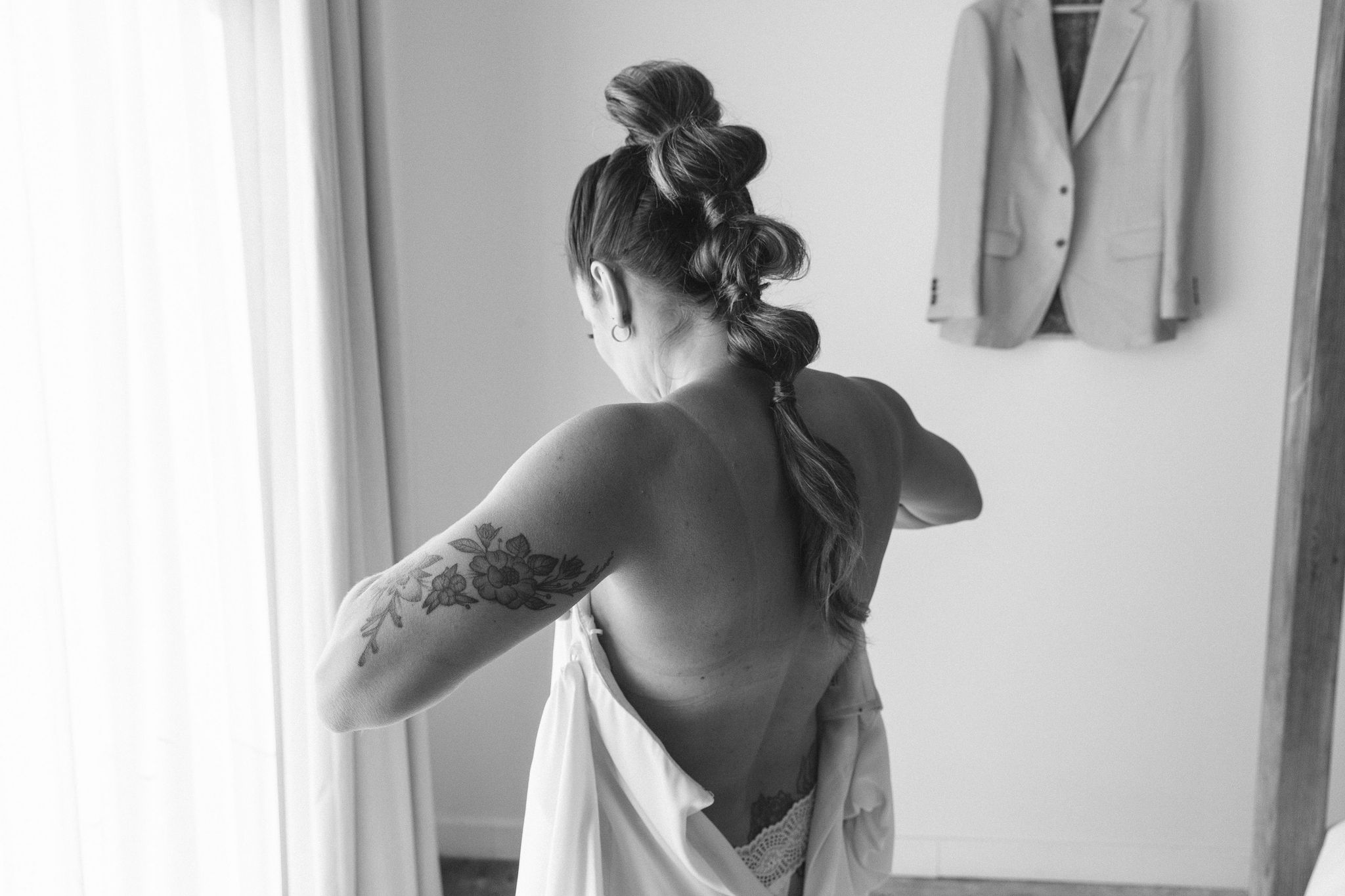 black and white style photo. bubble mohawk style long high ponytail. view from bride putting dress on from the back. floral arm tattoo. bridal getting ready.  verona gown by kamperett
