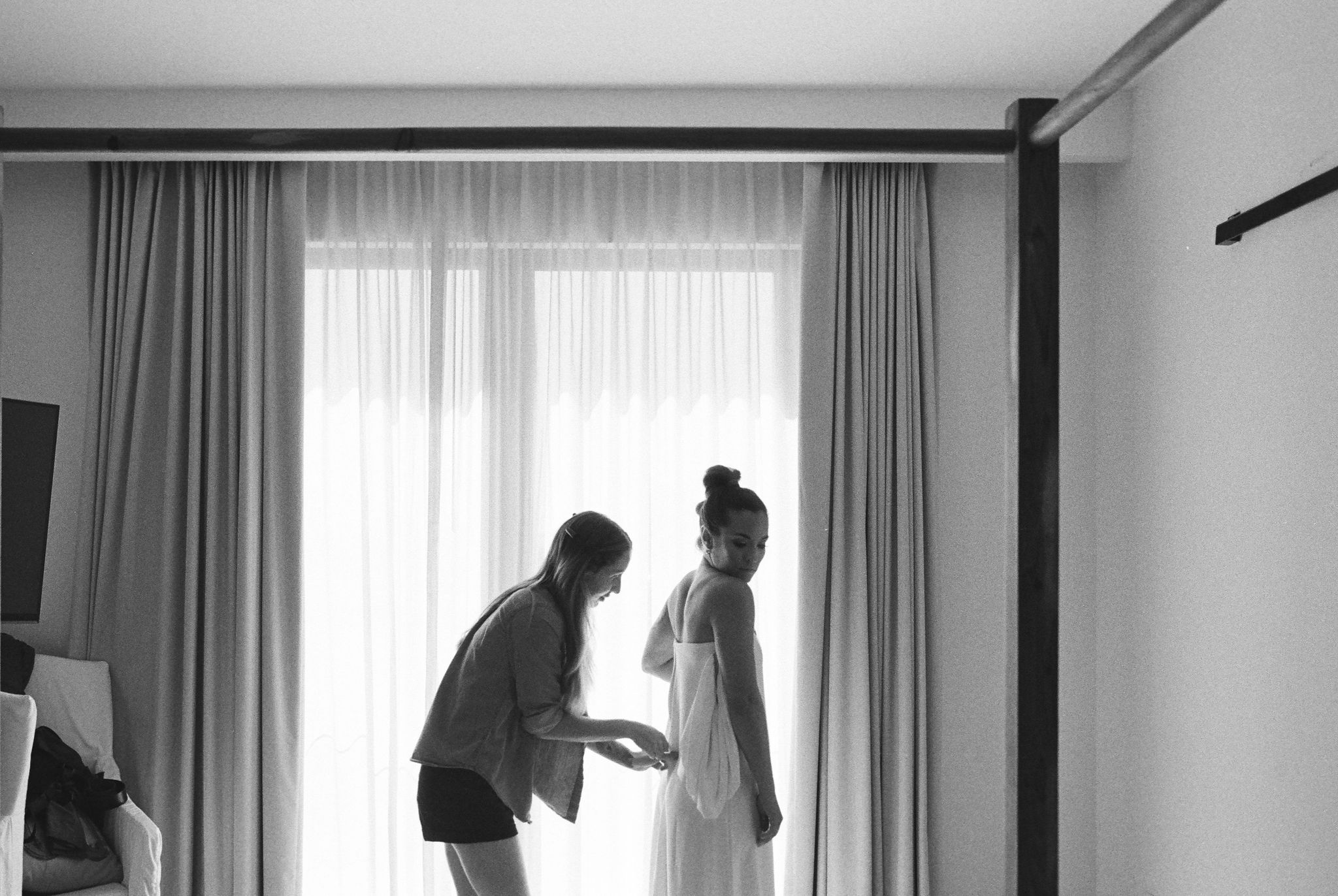 black and white photo of bride getting ready in front of window with a sheer curtain. bridesmaid zipping bride into dress. verona gown by kamperett