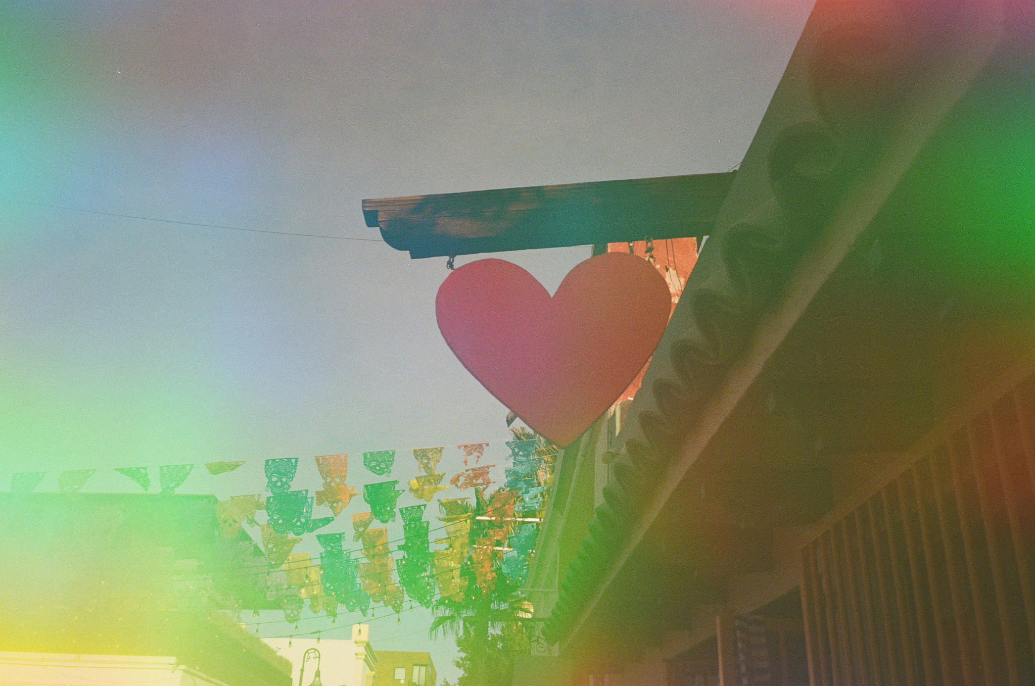 Close up of a wooden heart on streets of downtown San Jose Del Cabo, hanging outside of a shop on a wooden plank. In the background are colorful papel picado banners hanging over the street. The photo was taken on film with a grainy and iridescent sunflare finish.