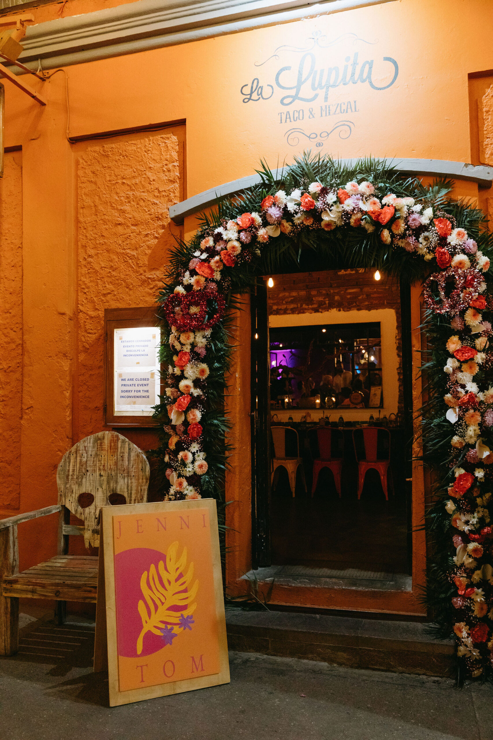 front of La Lupita restaurant San Jose Del Cabo, Mexico. Orange building with floral archway and distressed wooden chair with a skull-shaped back. wooden sandwich board with colorful (orange, pink, yellow and purple) design. Jenni and Tom welcome sign. 
