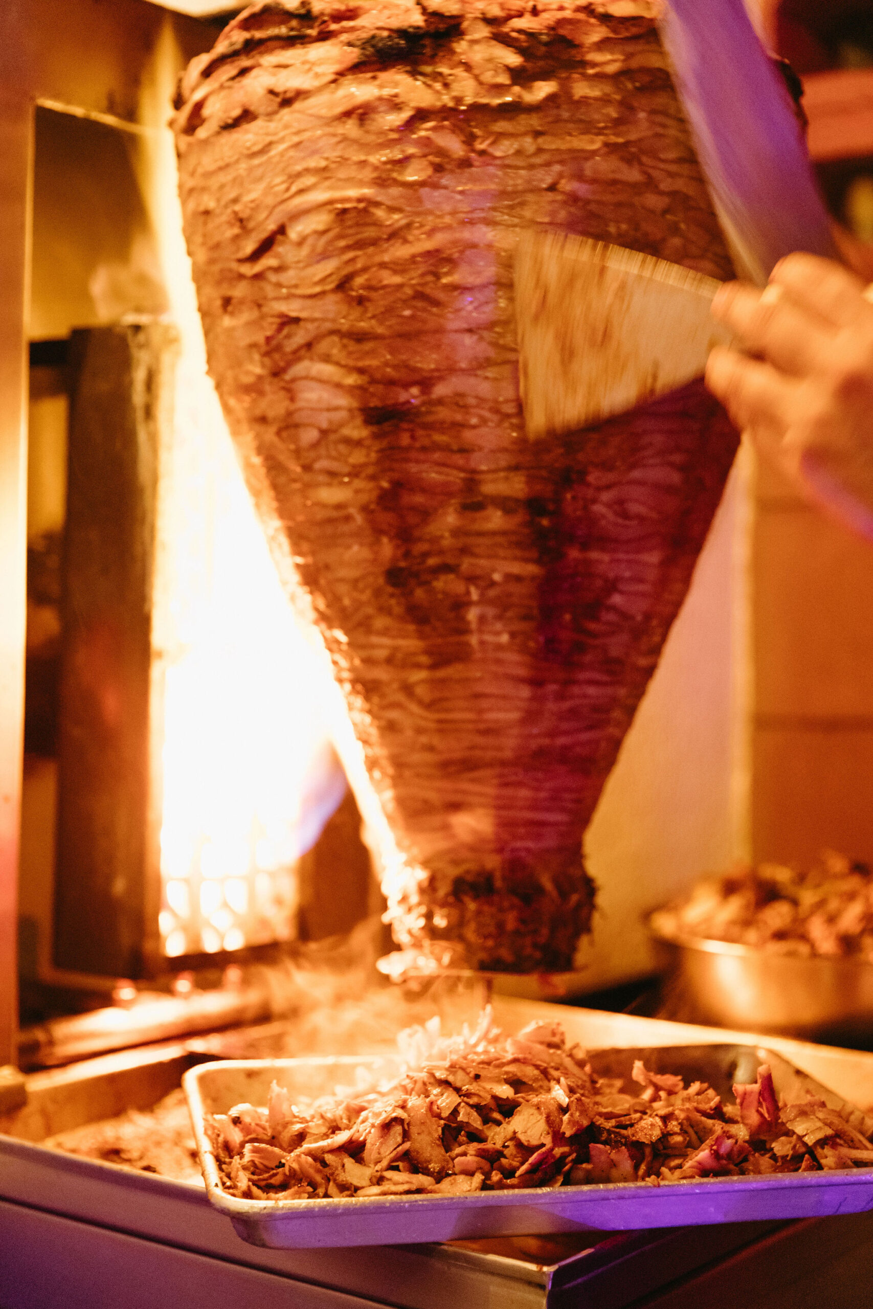 Al pastor on spicket and hand carving pork shavings onto tray at night with bright lighting from fire. 