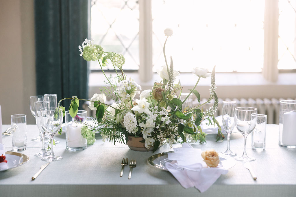 elegant white delicate florals with simple greens wedding table arrangements sage green linen tablecloth and white pillar candles clean clear glassware simple white napkin brushed gold flatware