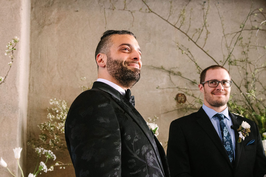 groom emotional aisle ceremony candid classy black suit with satin details dogwood tree branch ceremony alter delicate white florals and stone backdrop