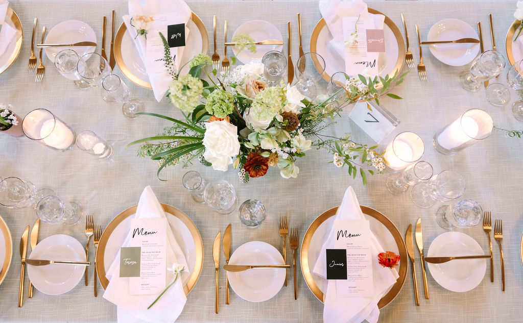 overhead view table top gold rimmed white chargers with gold flatware simple white napkin clean white and black name tags white pillar candles clean clear glassware delicate white florals and greens table arrangements 