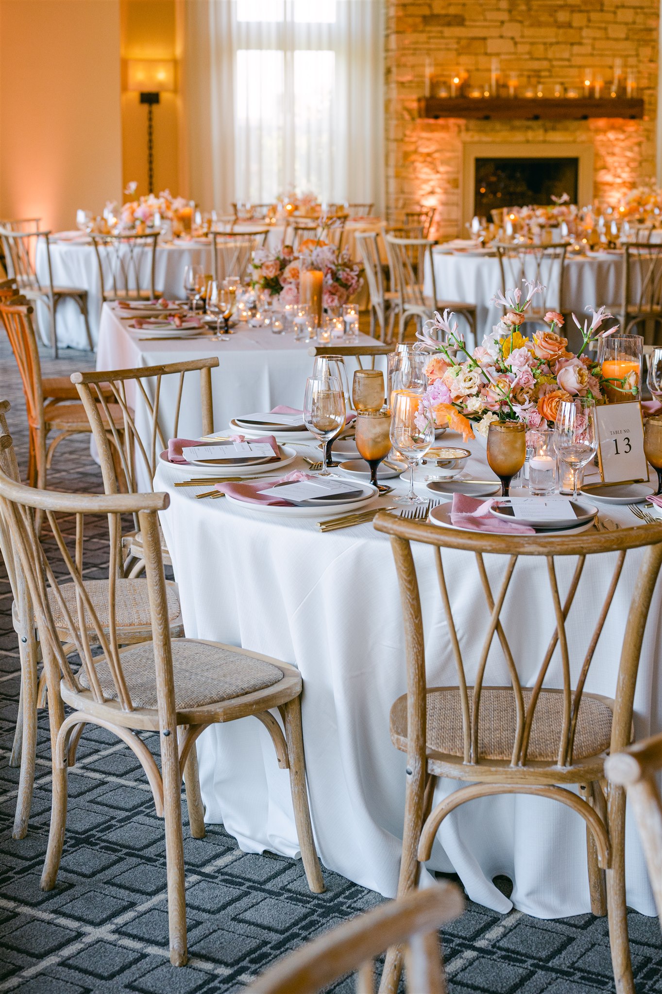 peach pink coral white delicate floral arrangement coral pillar candles clear glass candle votives with gold rim white clean table cloth simple stone dinner plates wedding reception wooden and rattan dining chairs gold framed simple table numbers smoked amber glassware pink napkin