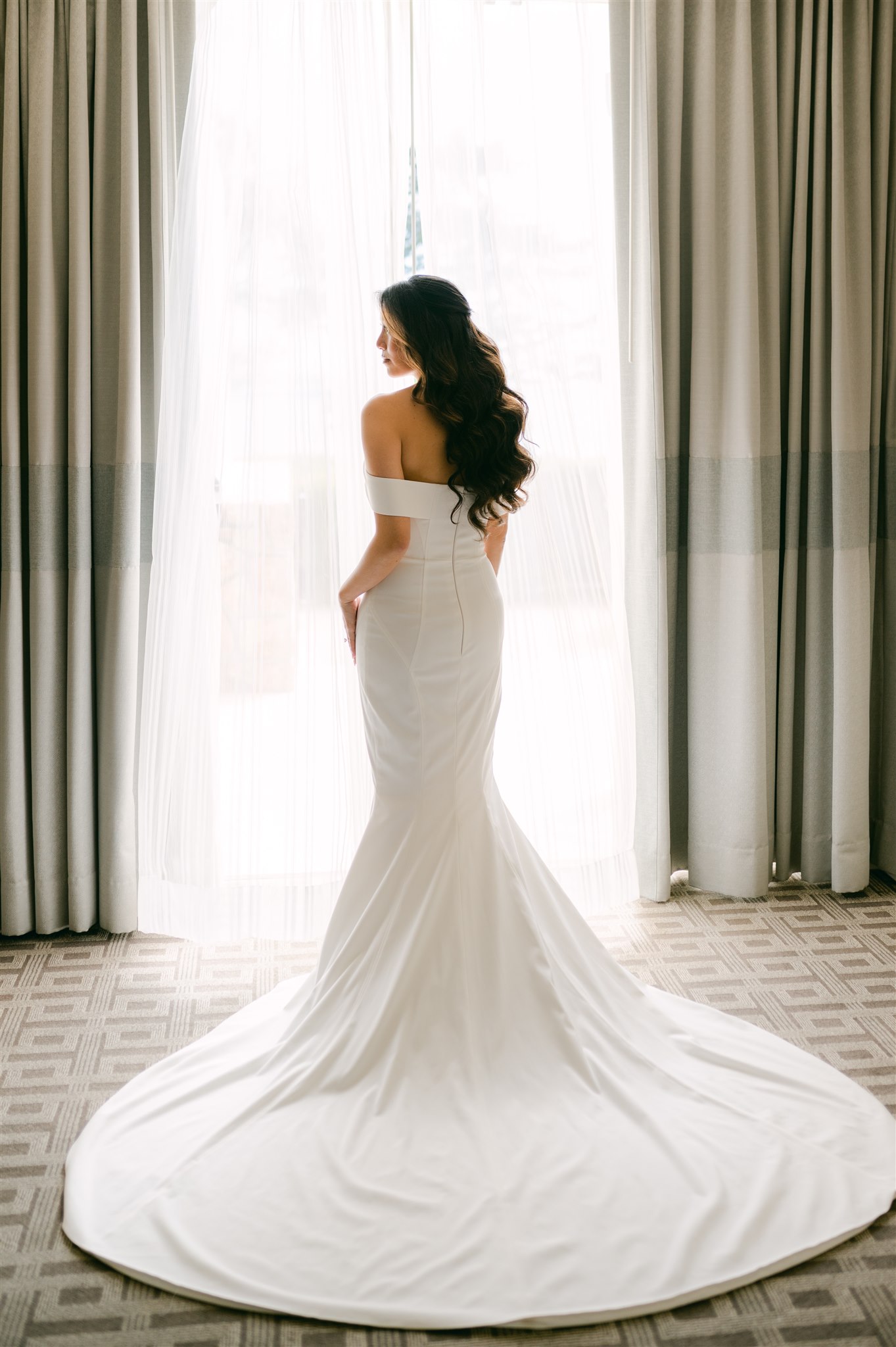 bride beach waves mermaid hair hour glass form fitting dress clean dress with off the shoulder details bride posing in front of backlit open window big train wedding dress